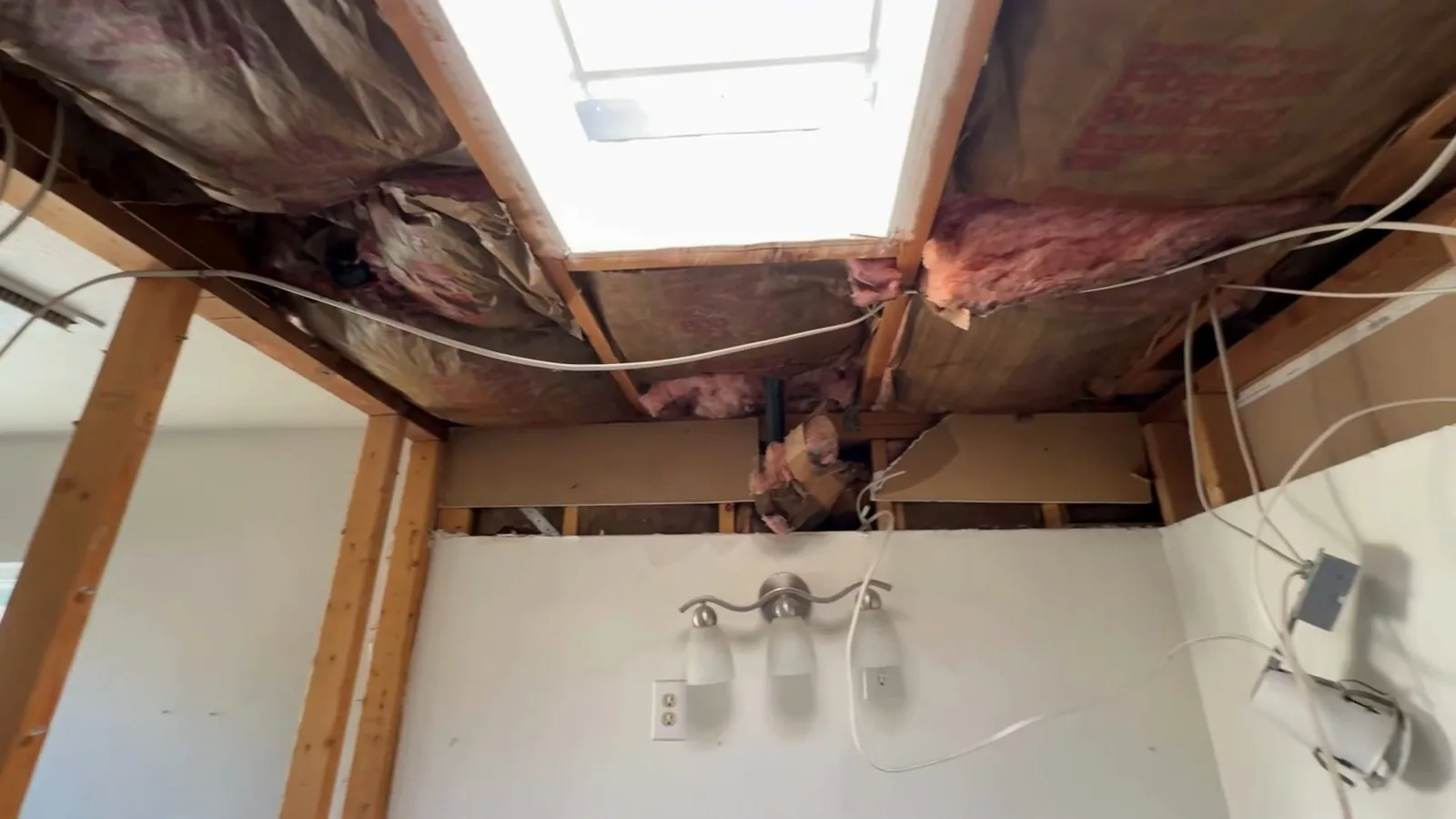 A bathroom ceiling opened up to bare joists and insulation, above two light fixtures on the wall.