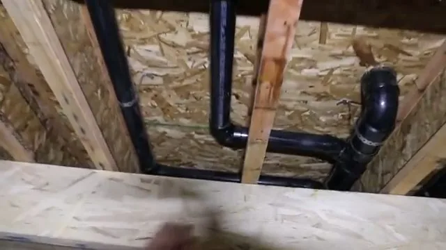 Black plastic drain pipes with elbows running through wood framing under a subfloor.