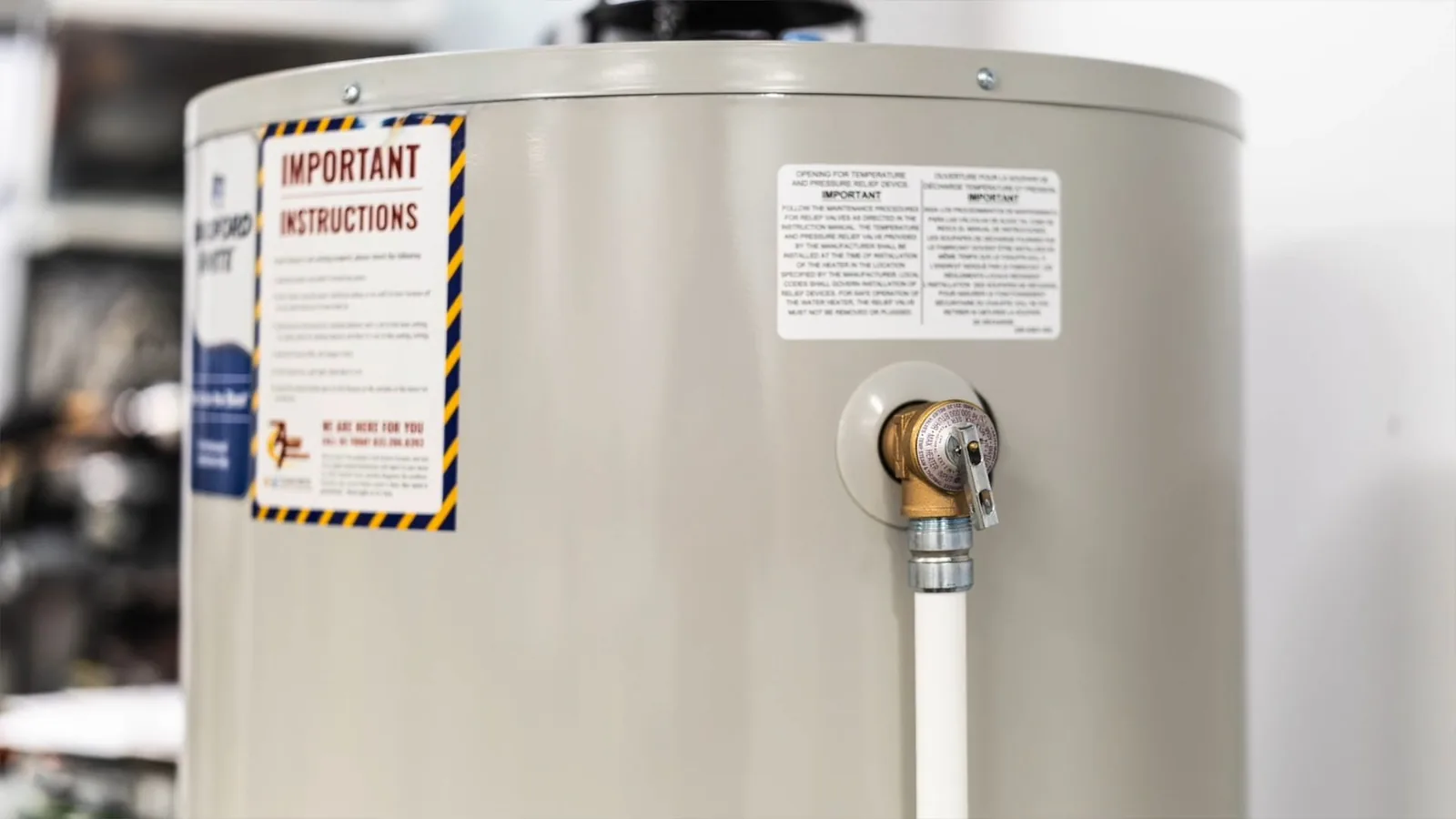 The side of a water heater tank with a brass relief valve and a white pipe running down from it, under a yellow warning label.