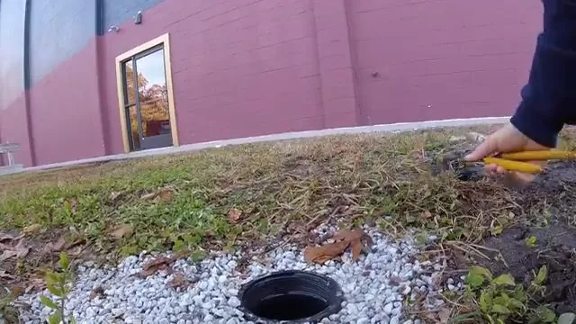 A hand setting a camera cable into a black sewer cleanout in a bed of white gravel beside a building lawn.