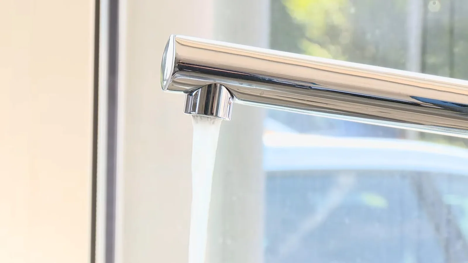 A chrome kitchen faucet with water running from the spout, with a window behind it.