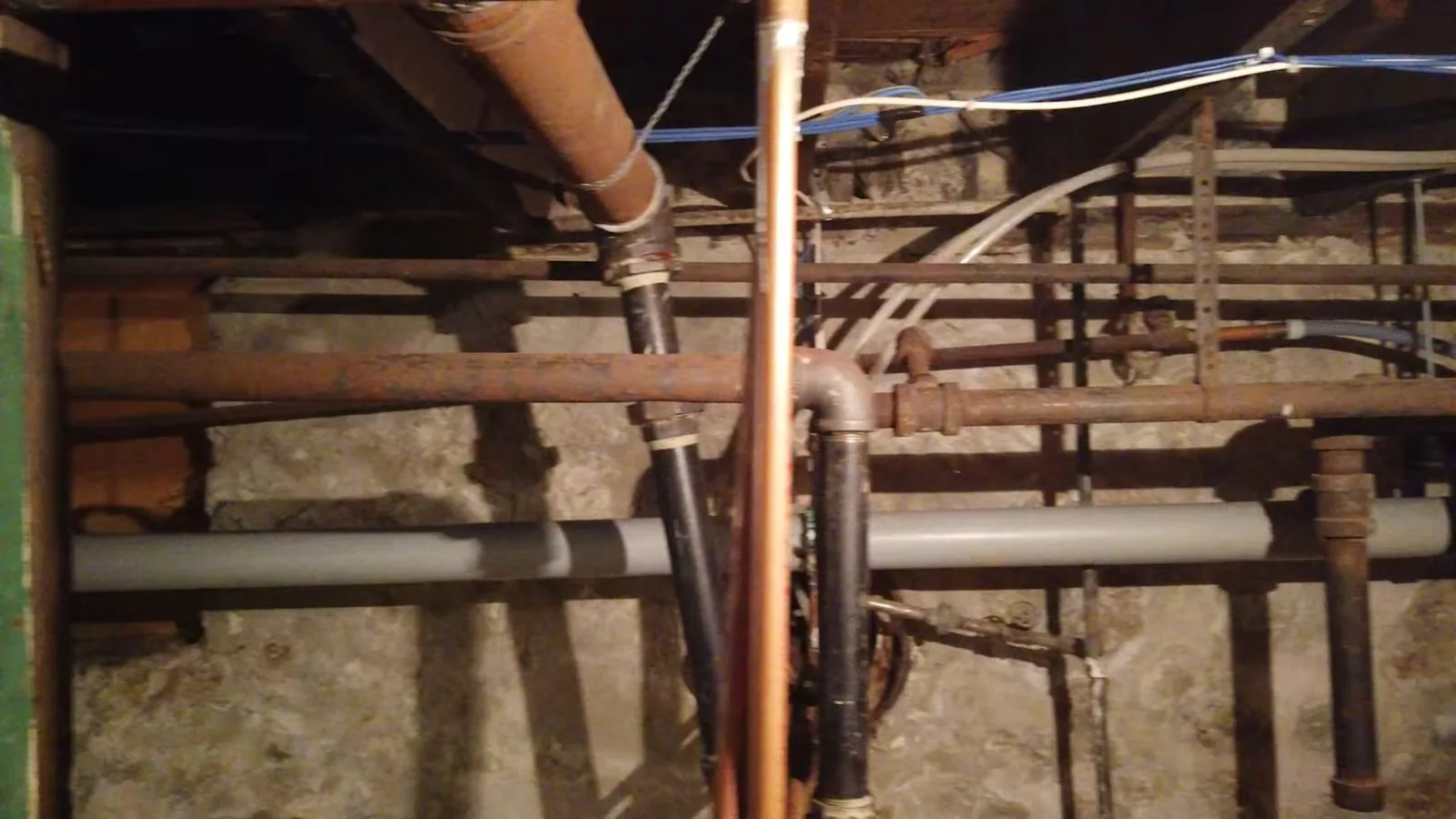 Copper, iron and white plastic pipes running under floor joists beside a stone foundation wall.