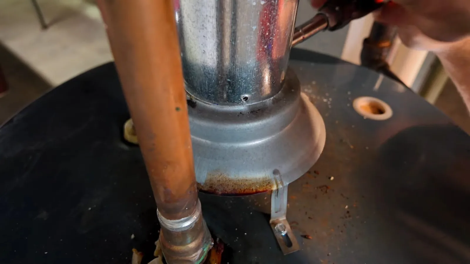 A copper pipe and a metal flue rising from the top of a black water heater, with rust around the base of the flue.