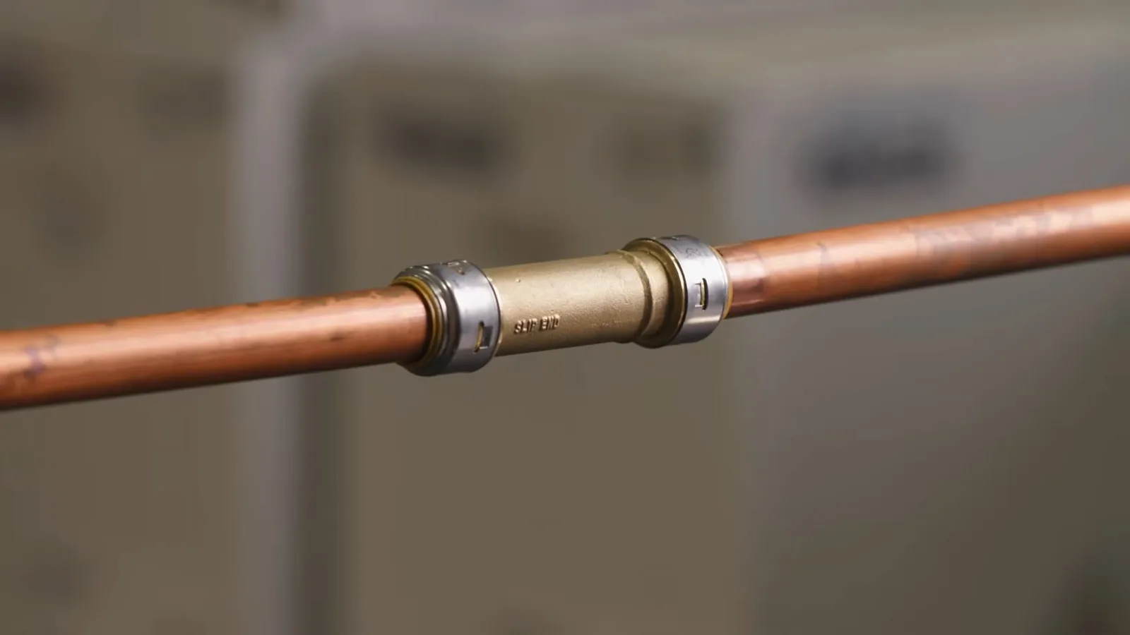 A copper water pipe joined by a brass coupling with steel grip rings, against a blurred background.