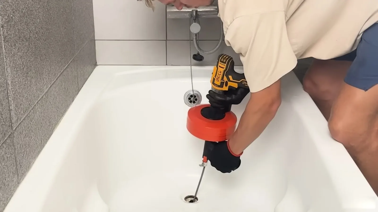 A person leaning into a white bathtub with a drill-driven drain snake feeding a cable into the drain.