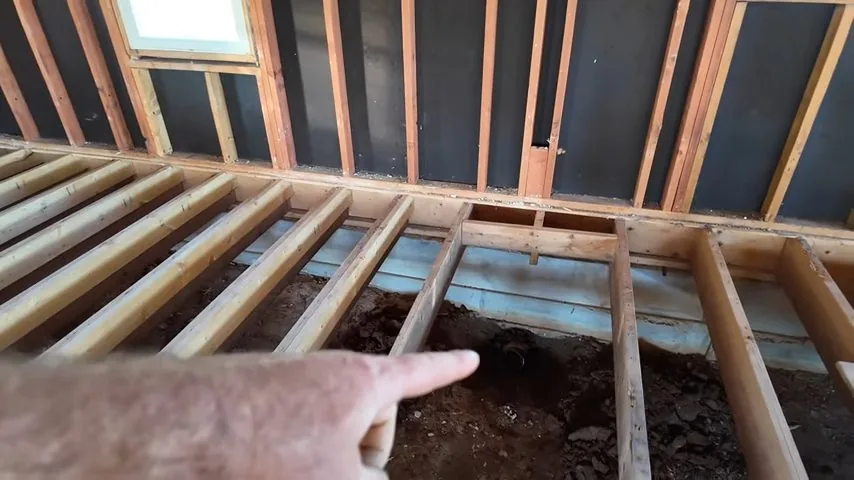 A floor opened down to wooden joists and subfloor, with a hand pointing at a spot between them.