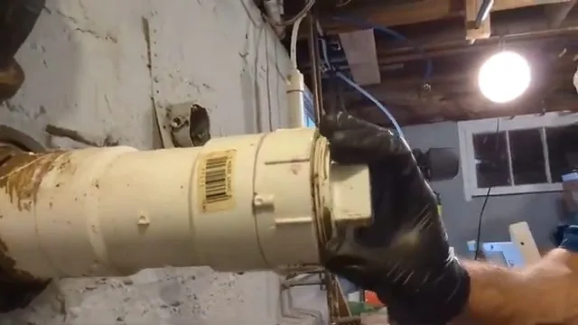 A gloved hand unscrewing the cap on a cream-colored sewer cleanout pipe in a basement.