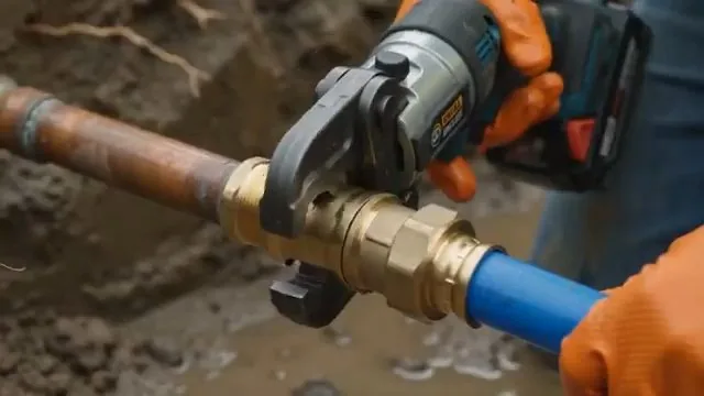Orange-gloved hands using a battery tool on a brass fitting that joins a blue PEX line to a copper pipe.
