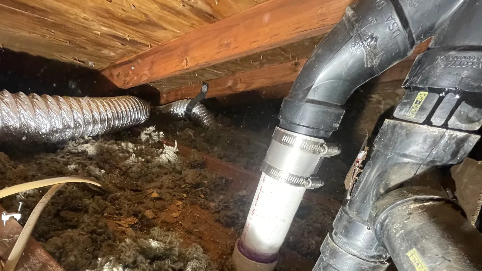 A gray drain pipe with a white coupling and metal clamps running under floor joists beside flexible duct.