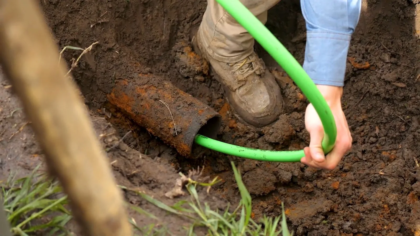 A gloved hand feeding a green liner tube into the open end of an old iron sewer pipe in a small dirt pit.