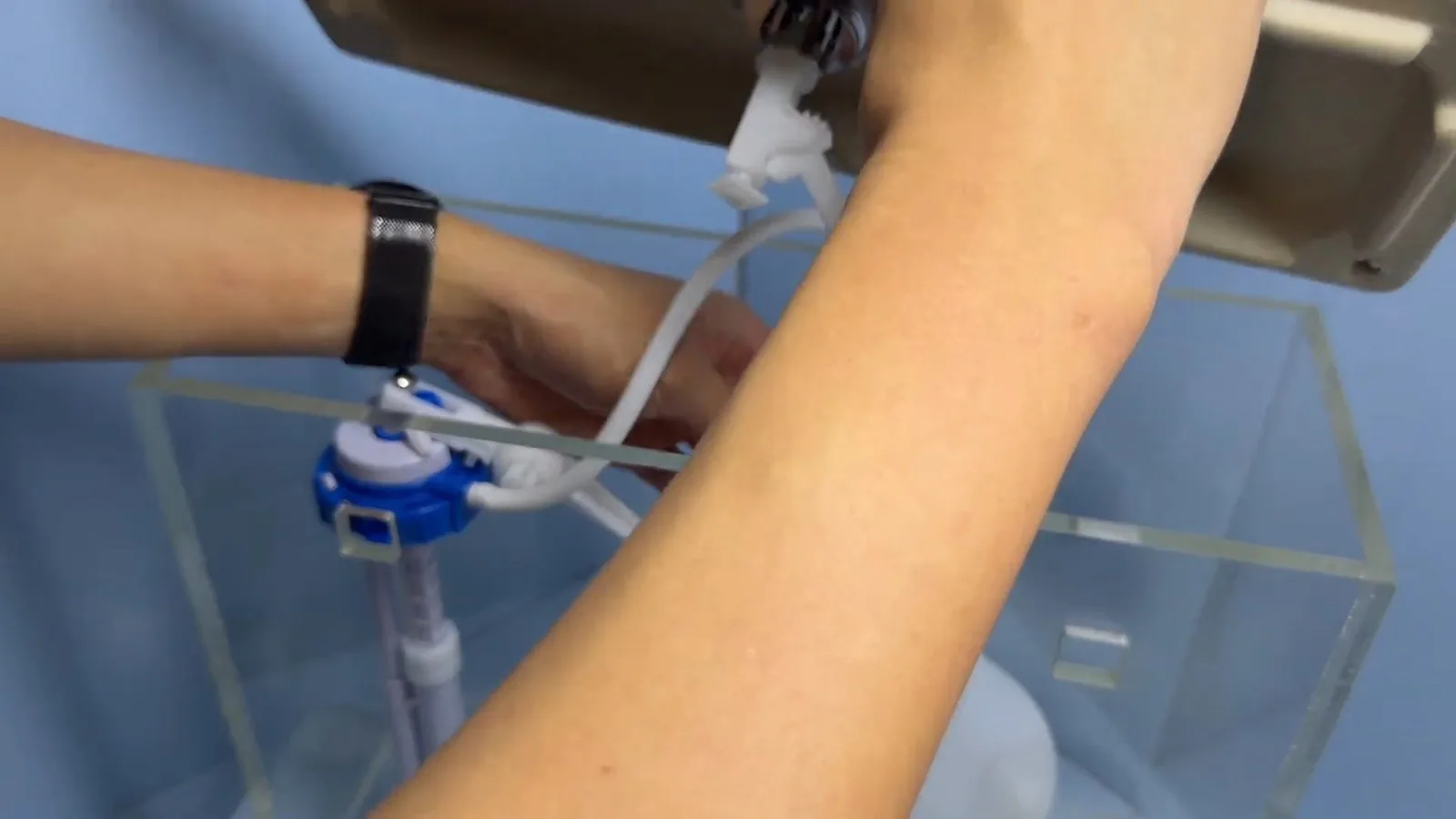A hand adjusting a blue fill valve inside a clear acrylic toilet tank.