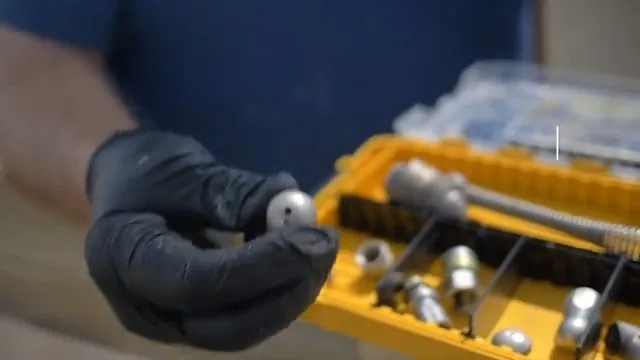 A gloved hand lifting a metal jetting nozzle out of a yellow organizer case that holds several other nozzles.