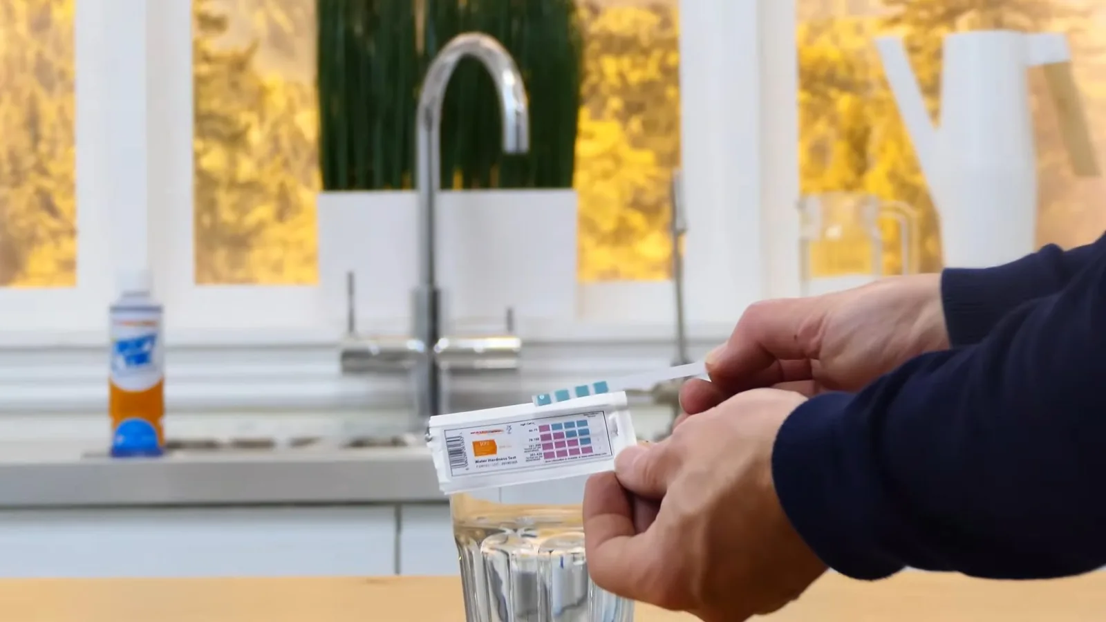 A hand holding a color test strip above a glass of water, with a chrome kitchen tap behind it.