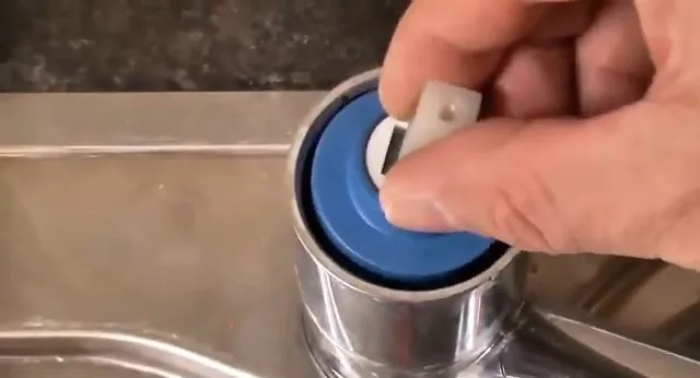 A hand lifting a blue cartridge cap out of the body of a chrome kitchen faucet.