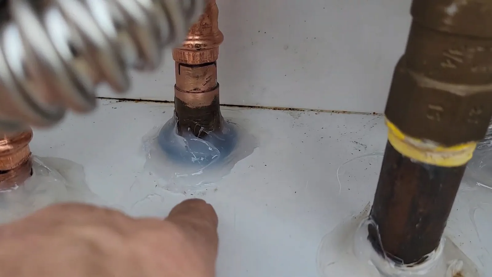 A hand pointing at a copper fitting sealed with blue compound next to a brass pipe on top of a white water heater.