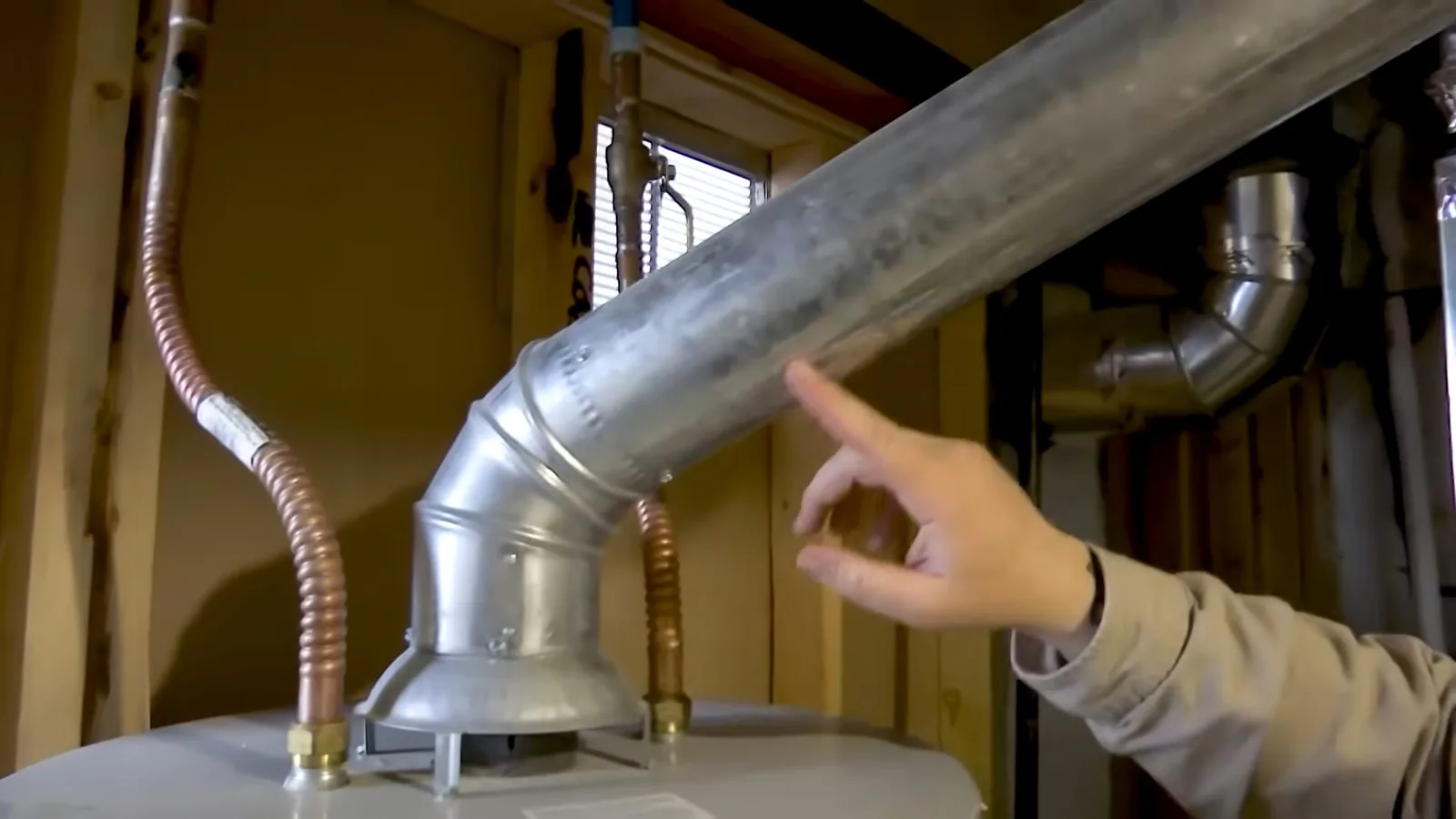 A hand pointing at a metal flue pipe above a water heater, with two flexible lines running up the wall.