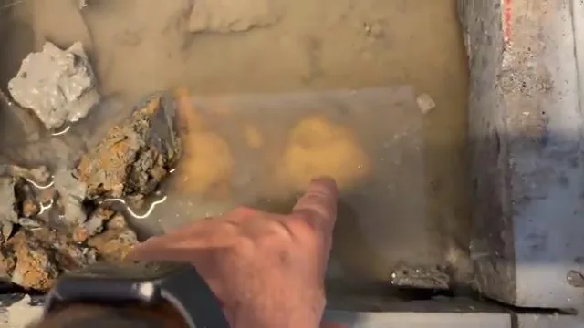 A hand with a watch pointing at brown water pooled in an opening beside the edge of a concrete slab.