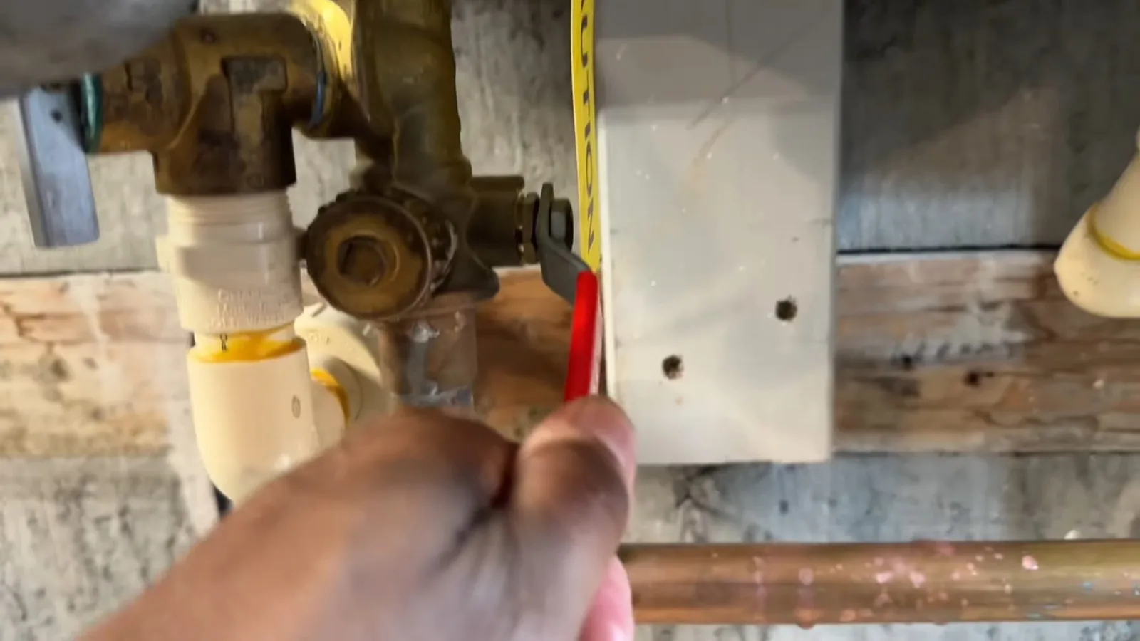 A hand reaching toward brass valves and white pipe fittings on a wall, with a measuring tape held beside them.