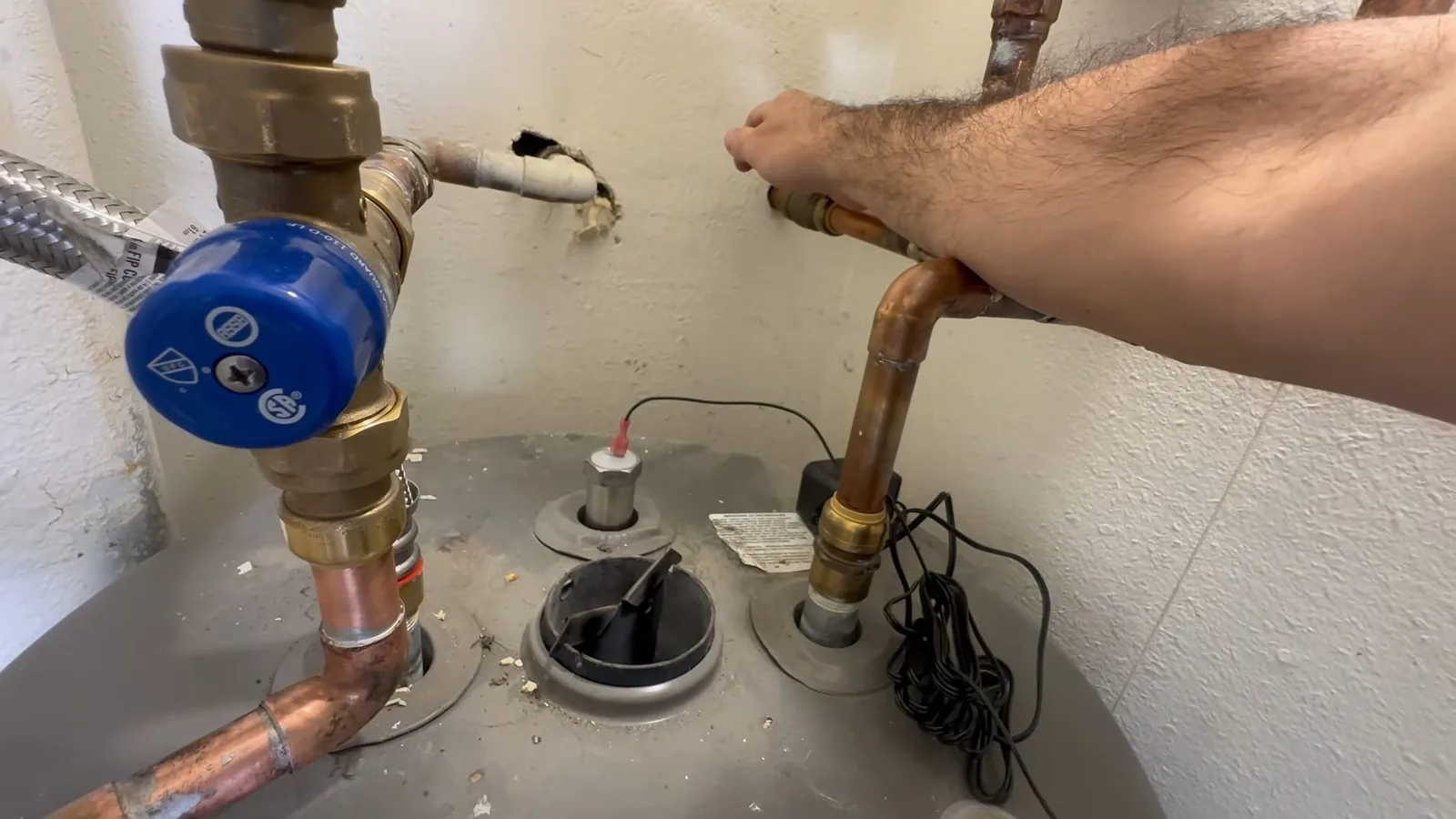 The top of a water heater tank with copper supply lines, a blue valve and an arm reaching in to a fitting.