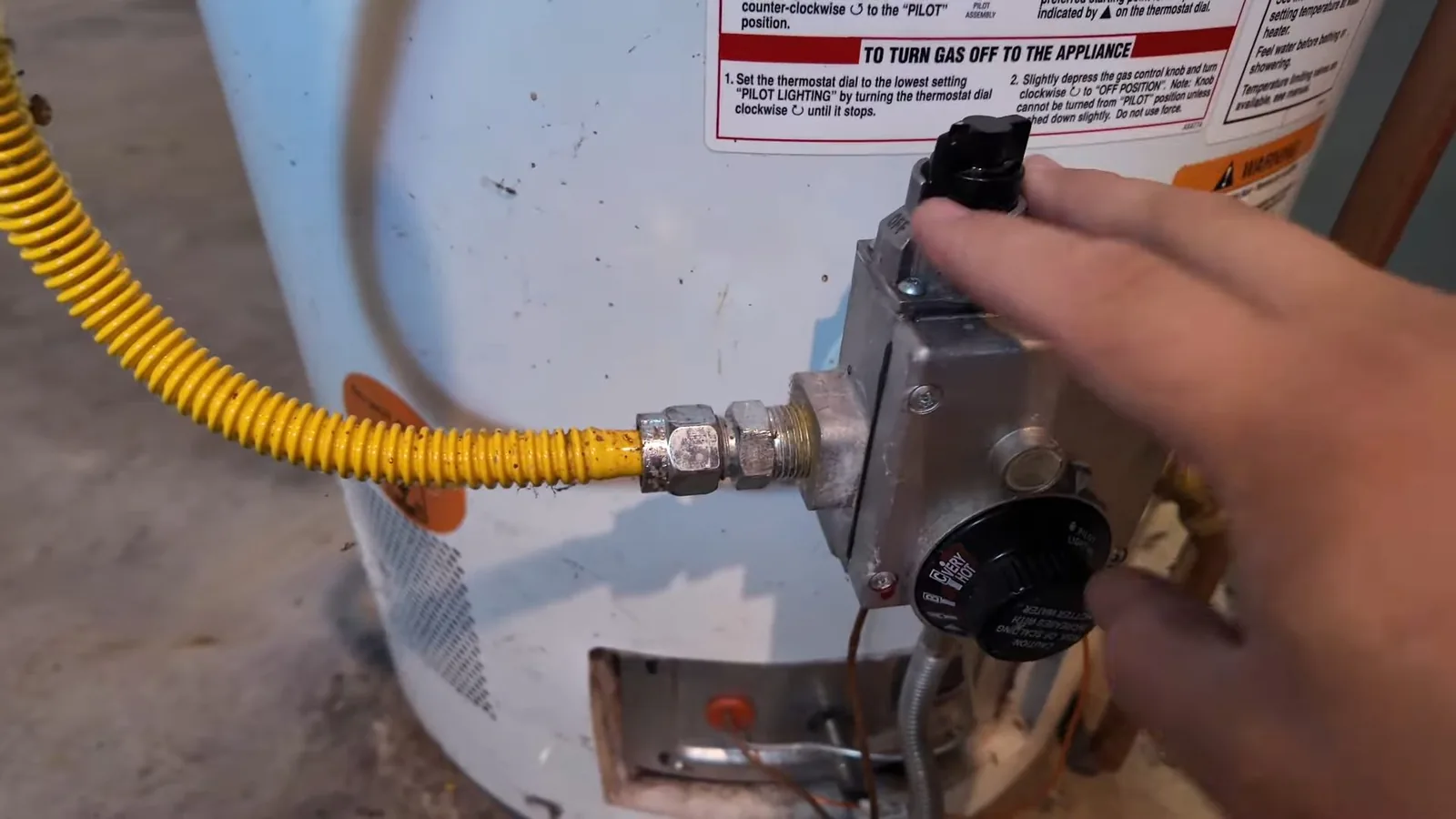 A hand turning the gas control knob on a water heater, with a yellow flexible gas line attached to the valve.