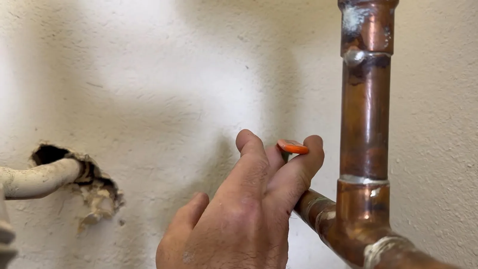 A hand holding an orange marker beside a copper pipe fitting next to a hole in a painted wall.