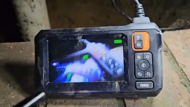 A handheld camera monitor with orange buttons showing the inside of a pipe on its screen.