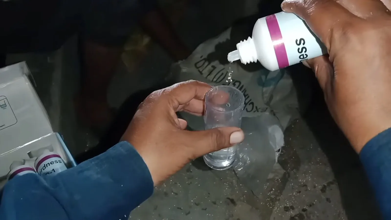Two hands holding a small bottle over a clear cup while test drops are added to the water.