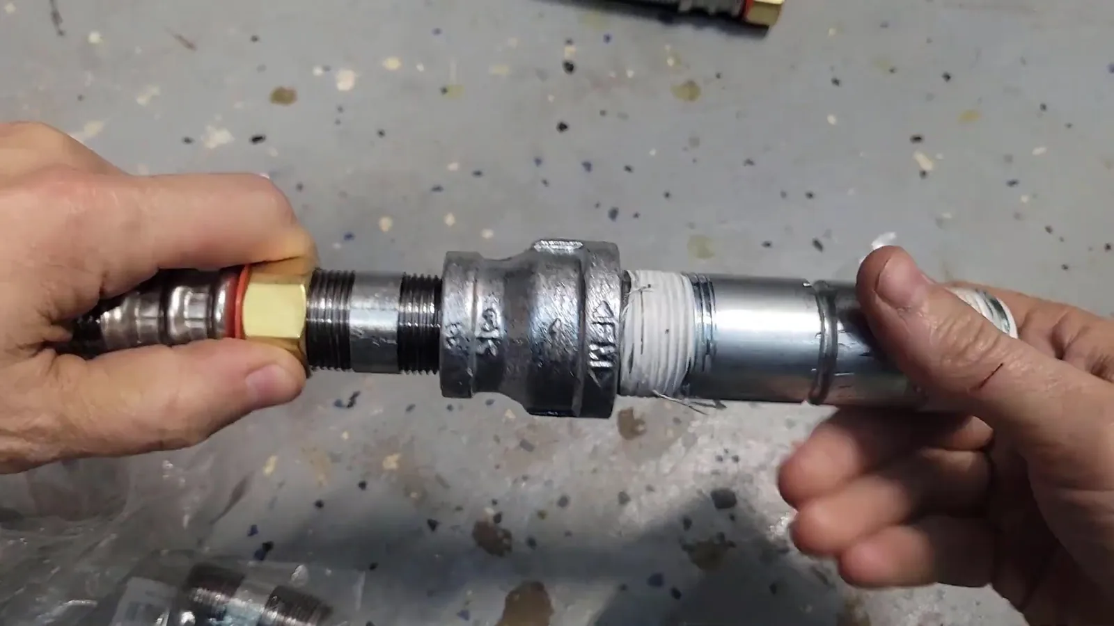 Two hands joining a galvanized union fitting to a brass connector over a speckled gray floor.