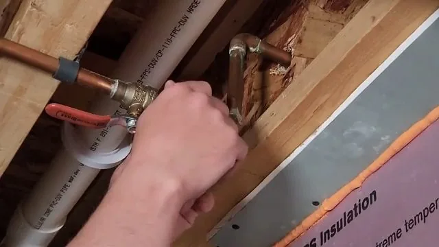 A hand fitting a copper connection beside a white pipe between wooden joists under a floor.