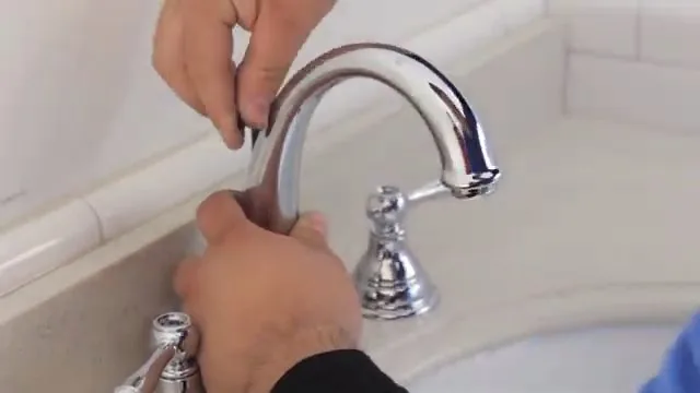 Two hands taking the handle off a chrome two-handle bathroom faucet over a white basin.