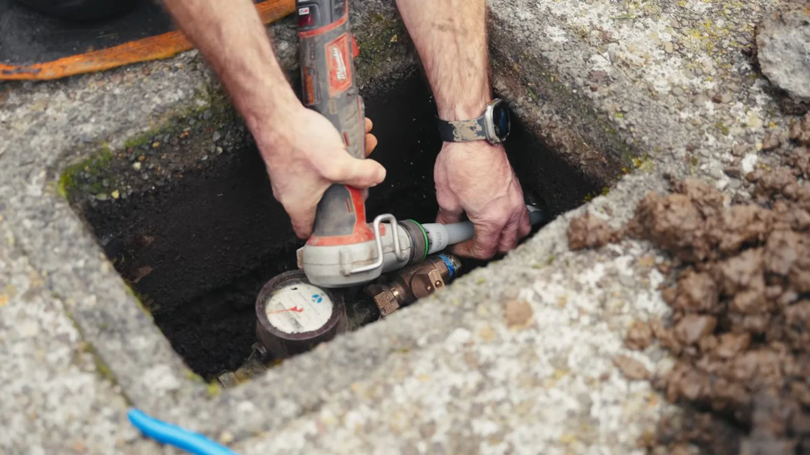 Two hands working on a water meter and its fittings inside a concrete ground box.