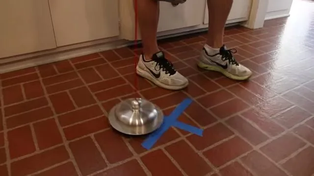 A metal listening disc on a tile floor next to a blue tape mark, with a person's feet standing beside it.
