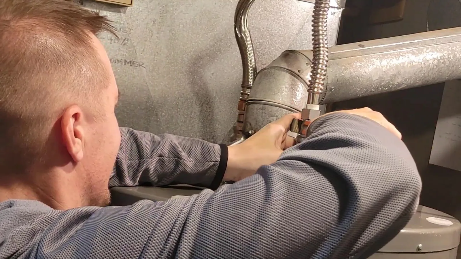 A man with short hair reaching in to connect a flexible supply line to the pipework beside a water heater.