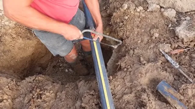 A man standing in a dirt pit beside a black pipe, holding a hacksaw and a tape measure against it.