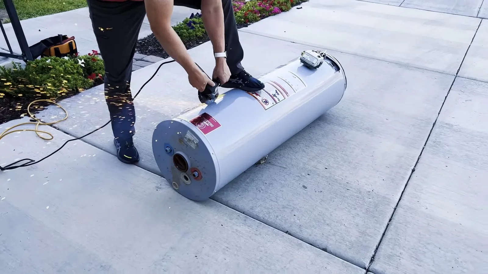 A new tank water heater lying on a concrete driveway with a person bent over it, working on the tank.