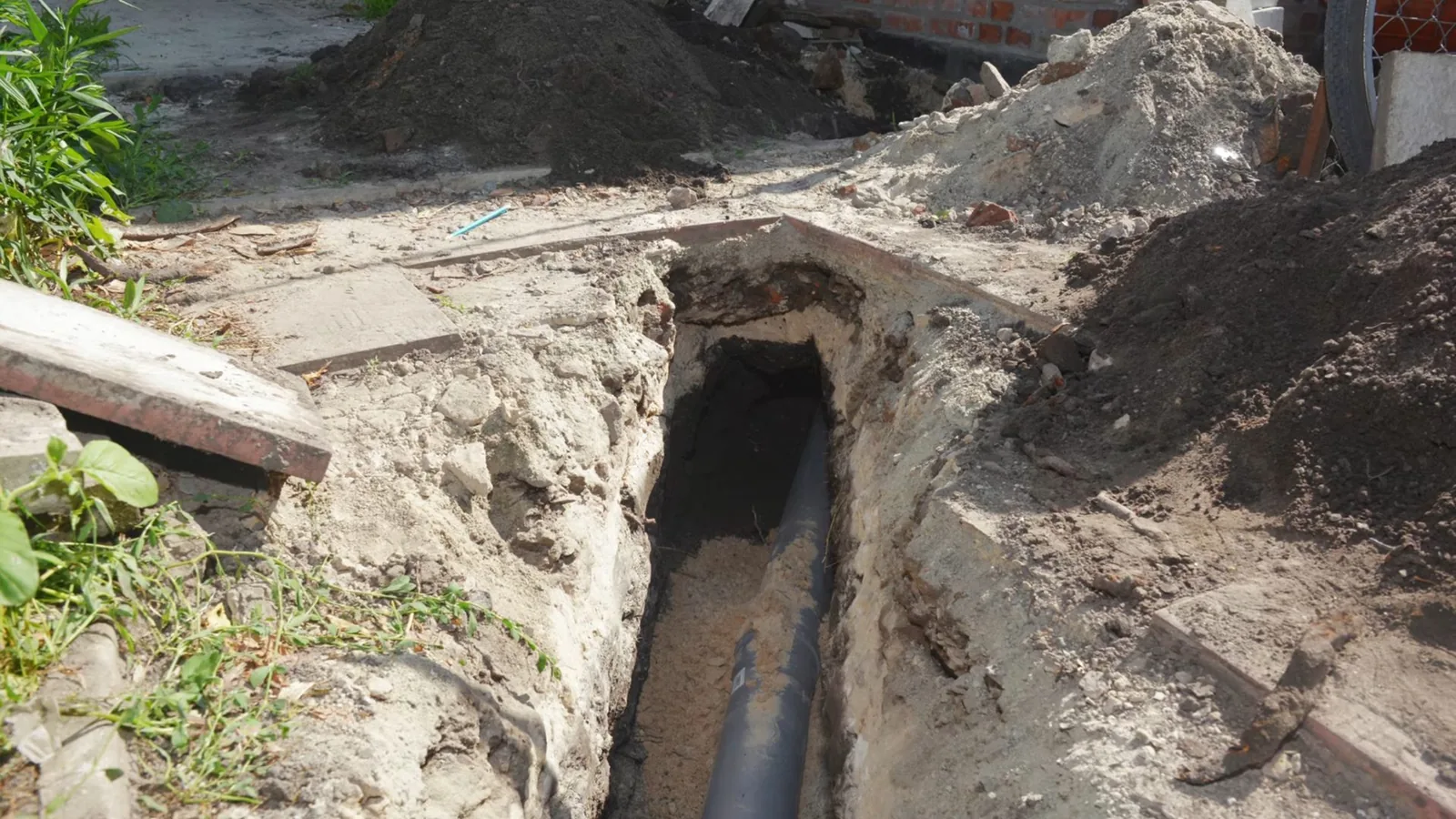 Open trench beside a house with a black sewer pipe running along its bottom.