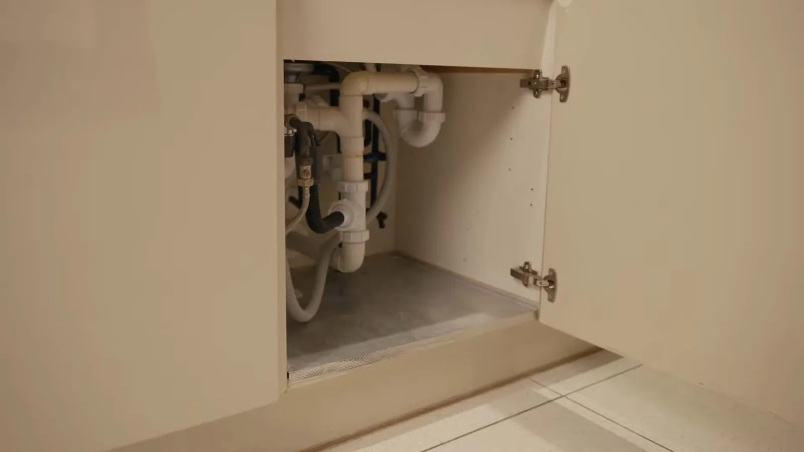 An open under-sink cabinet showing white drain pipes, a P-trap and supply lines above a gray floor mat.