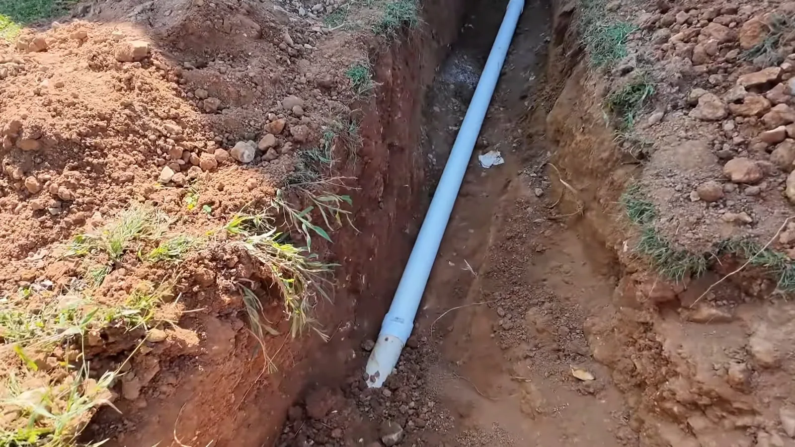 A trench cut through red clay and grass with a white pipe lying in the bottom.