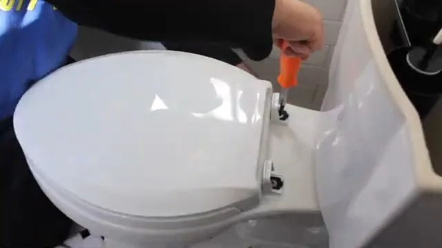 A plumber in a blue top working beside a toilet, with an orange screwdriver at the tank.