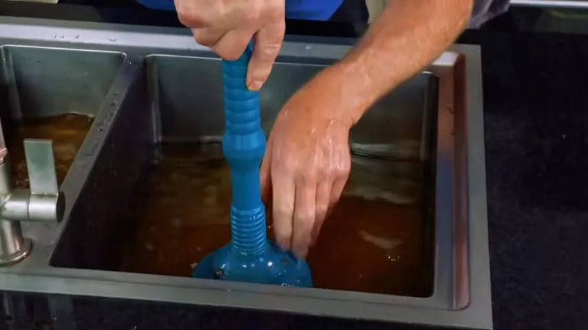 A hand pressing a blue plunger down in a double sink standing full of brown water.