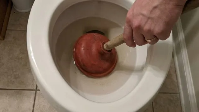 A hand pressing a red plunger down into a white toilet bowl.