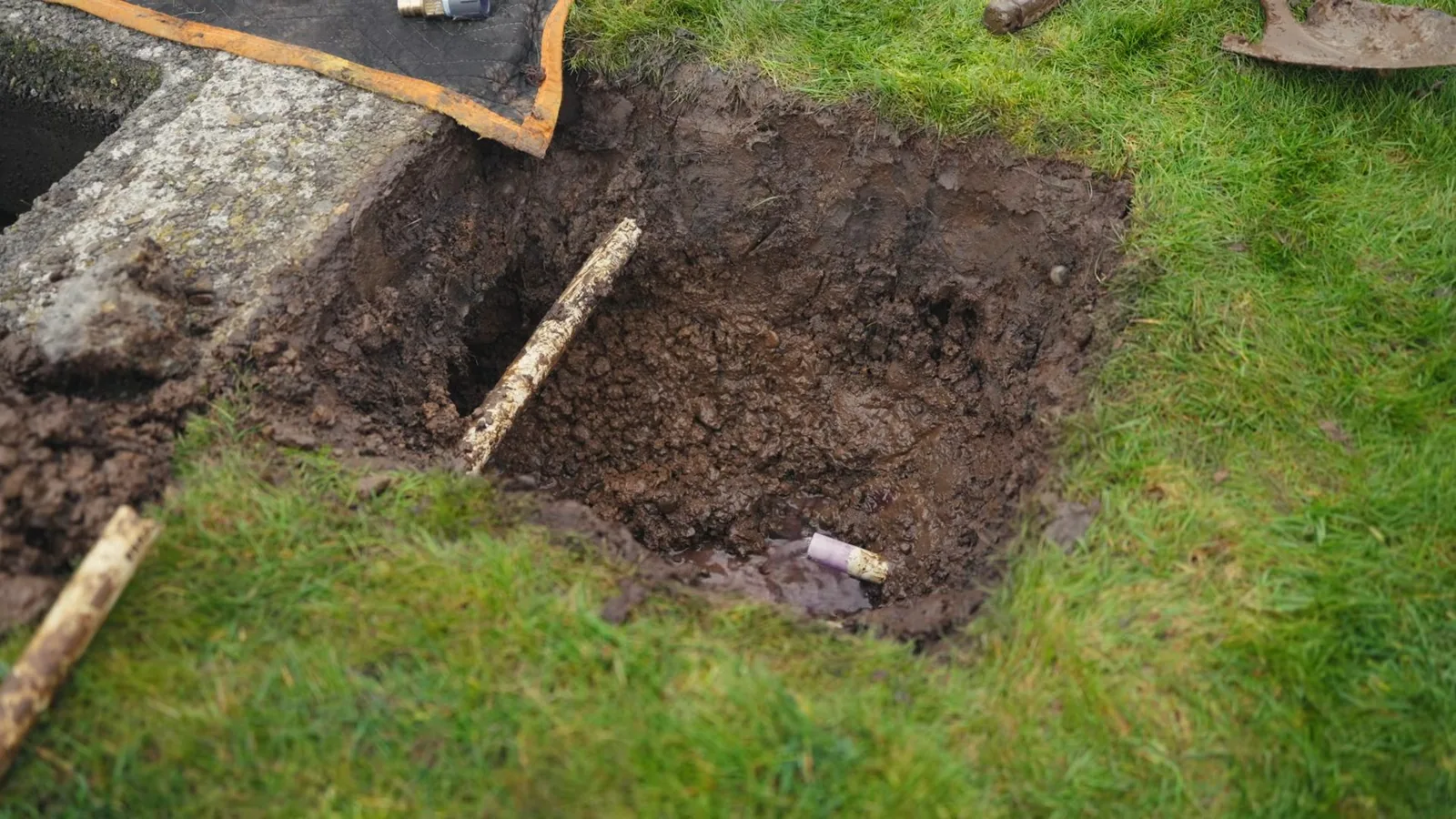 A square pit dug in a lawn with a shovel lying beside it and a pipe end visible at the bottom.