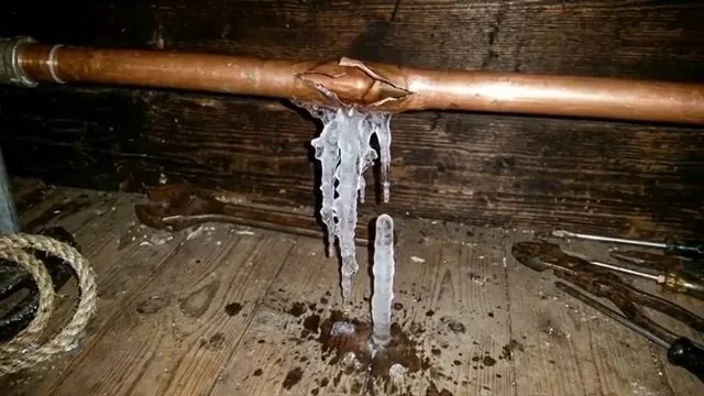 A split copper pipe running across a wooden wall with ice hanging from the burst and a wet floor beneath it.