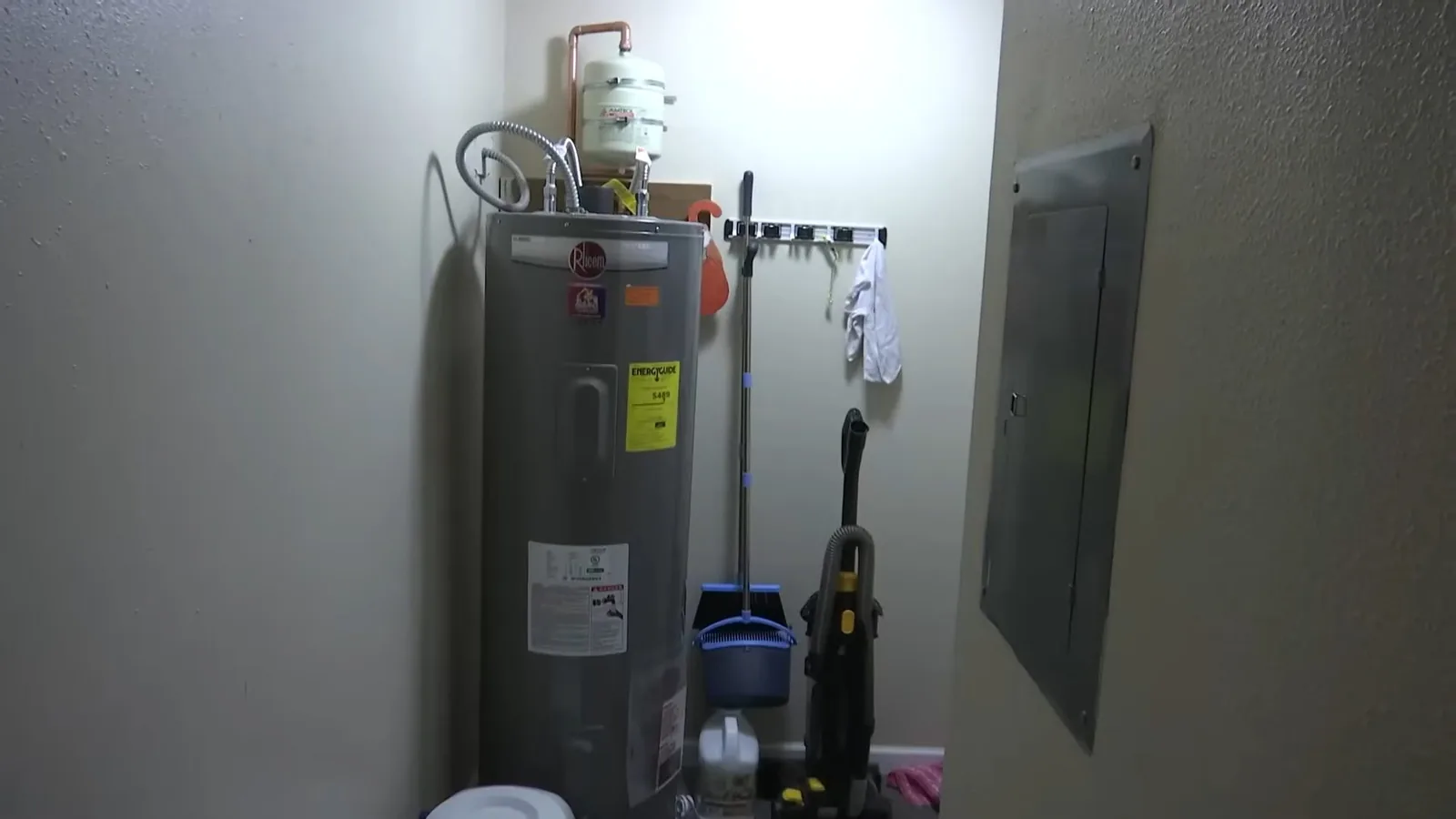 A tank water heater standing in a small utility room beside a mop, a bucket and a gray electrical panel.