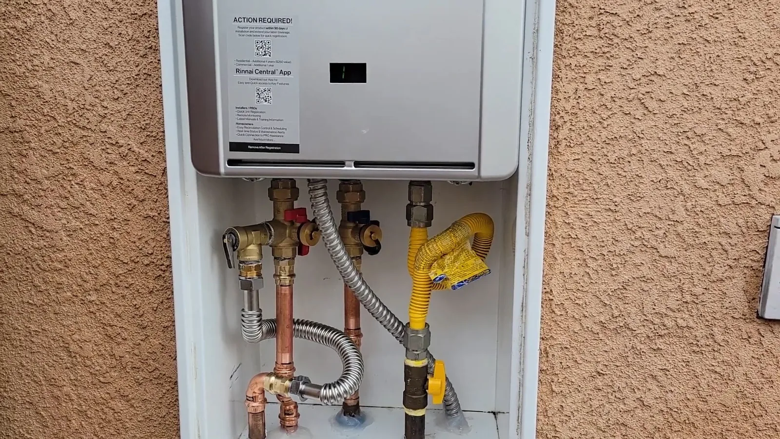 A tankless water heater mounted on a stucco wall with its pipes running underneath.