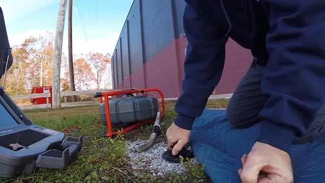 A technician kneeling beside a camera reel and case while feeding a camera line into a cleanout in the grass.