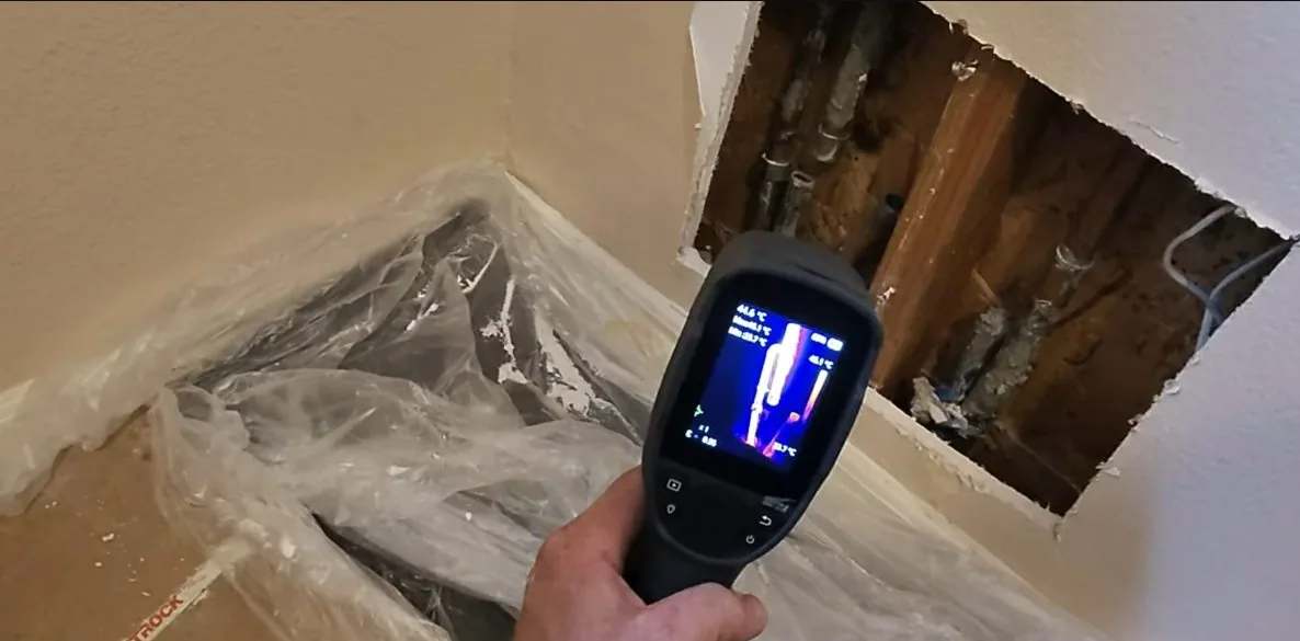 Handheld thermal imager held over an opened wall and floor corner, with a heat image on its screen.