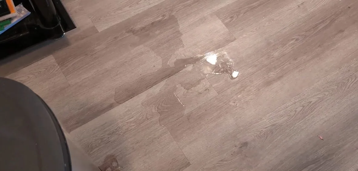 A small puddle of water on a wood-look floor beside a dark appliance.