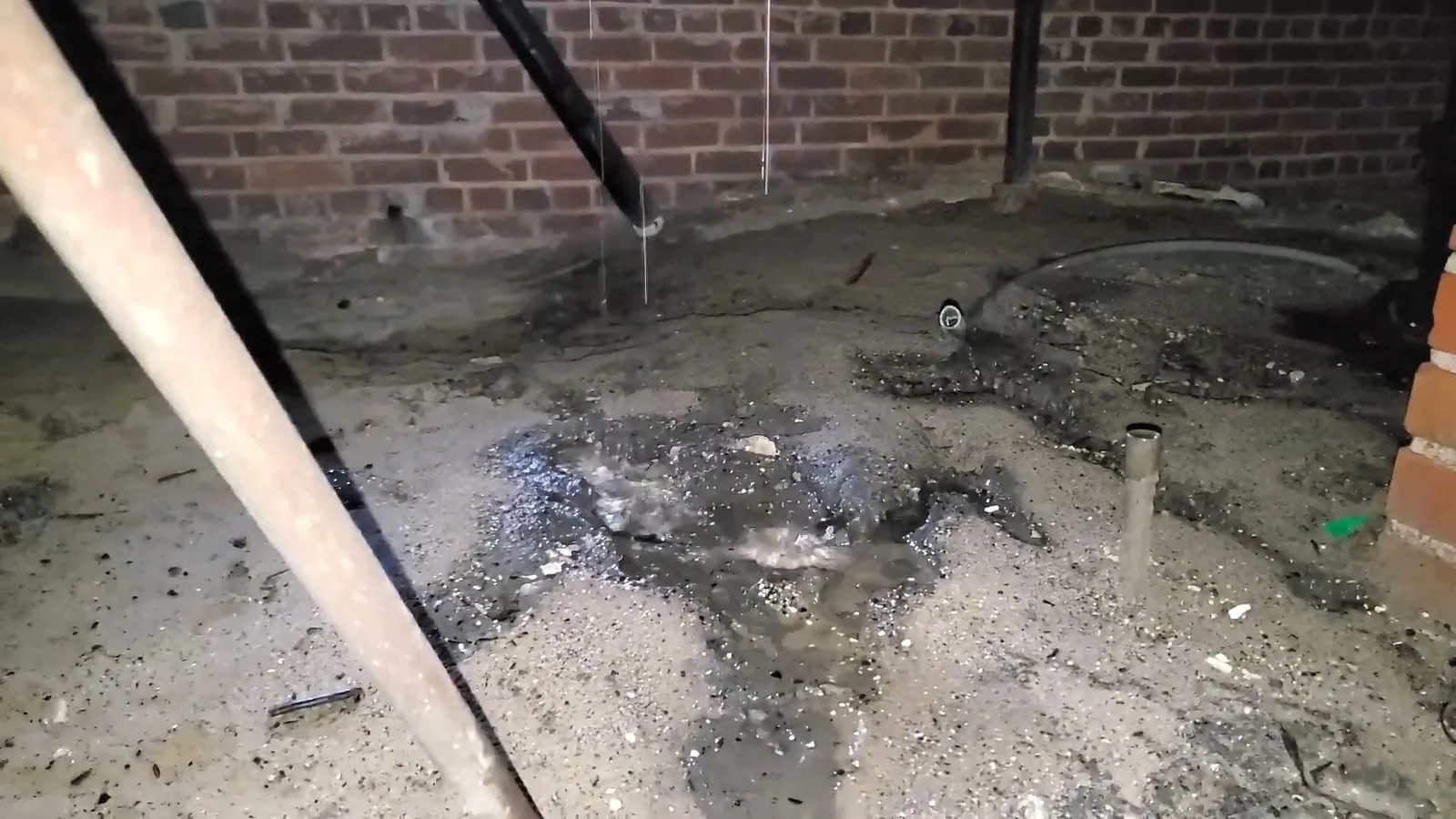 A wet crawlspace floor with a puddle of water spreading beneath a pipe and a brick wall behind it.