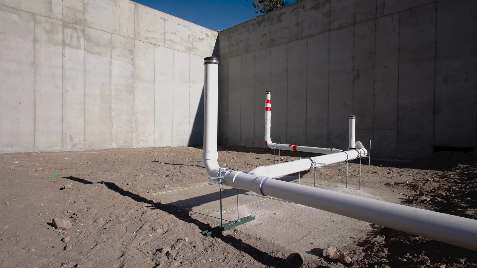 White plastic drain pipe with two vent risers lying on bare ground beside a tall concrete wall.
